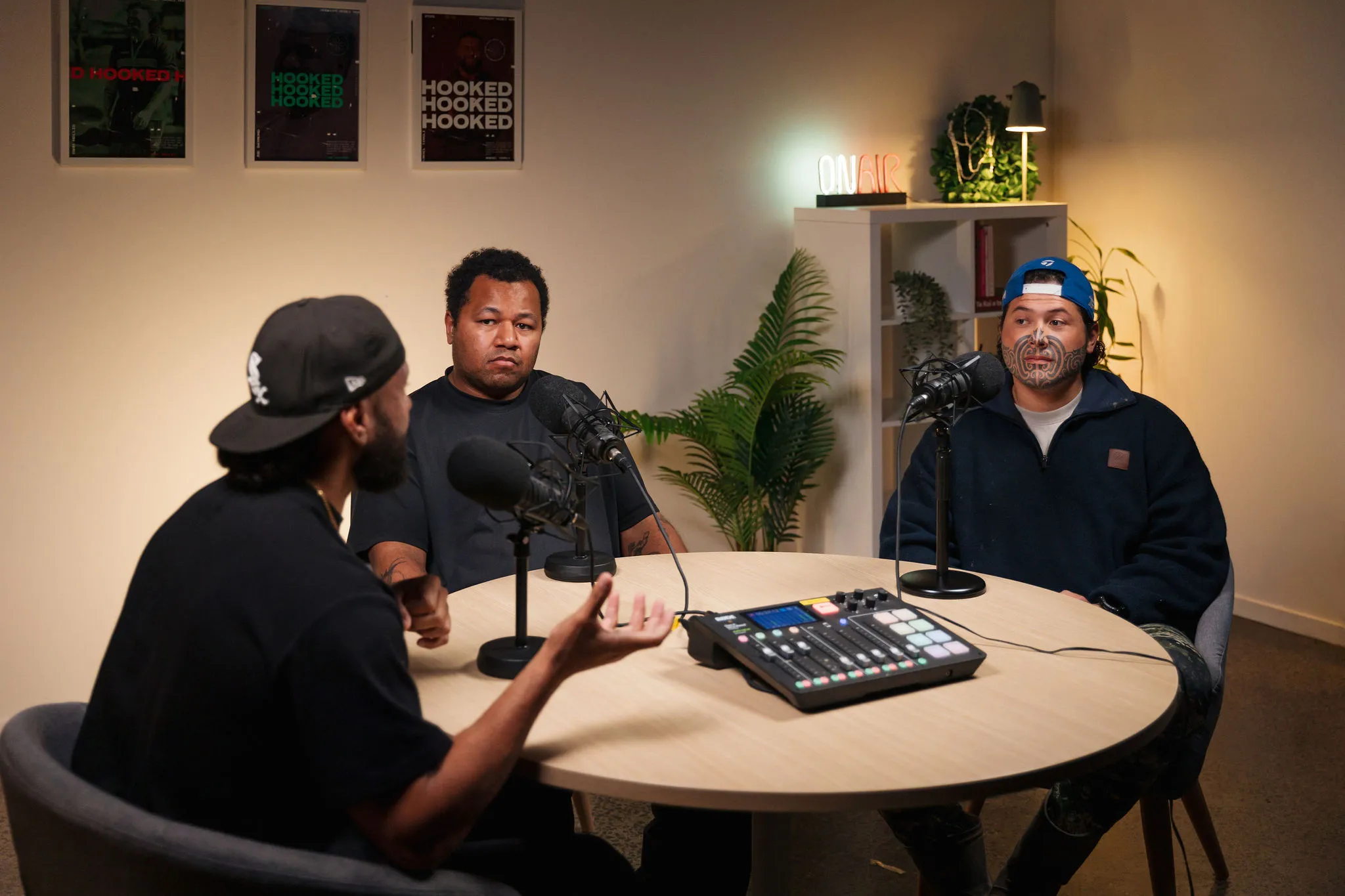 The Hooked podcast set: Joe Daymond, Montel Tivoli and Tahu Hollis in kōrero around the studio table, the show’s posters and the ON AIR sign behind them.